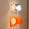 Two modern wall sconces mounted on a beige wall, one in silver with a warm yellow globe and one in orange with a white globe, both turned on.
