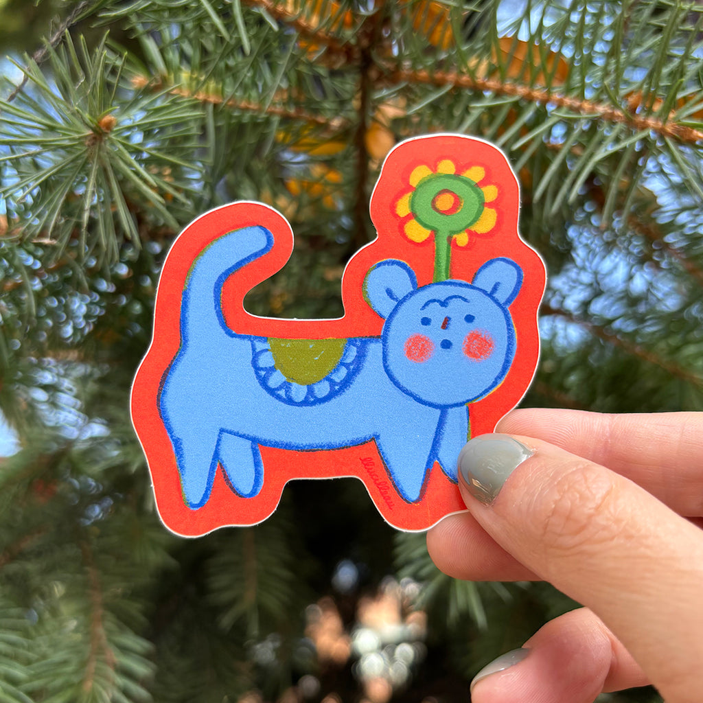 Colorful sticker of a blue creature with a red border held in front of green foliage