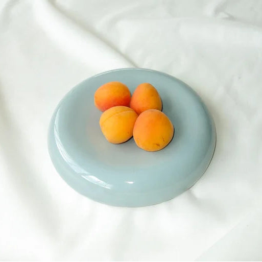 A round, shallow light blue dish holding four apricots, set against a backdrop of draped white fabric.