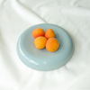 A round, shallow light blue dish holding four apricots, set against a backdrop of draped white fabric.
