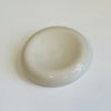 A round, shallow ceramic dish or saucer in a light beige or off-white glaze, photographed from a top-down angle on a plain white surface.