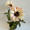 A bouquet of artificial zinnia flowers with cream-colored petals speckled with pink and white, dark brown centers, and green leaves, arranged in a small pot against a plain white wall.