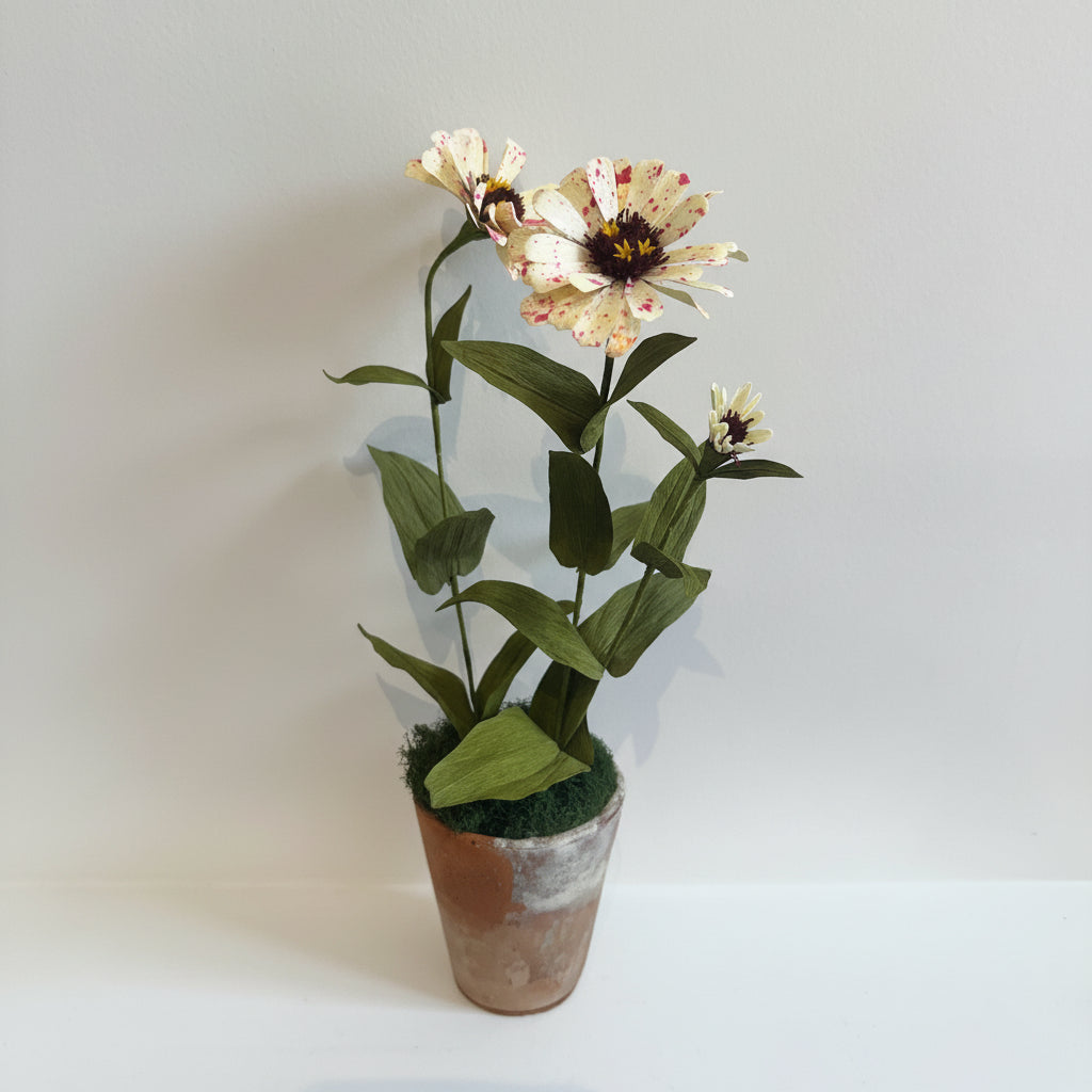 A potted artificial flower arrangement featuring three cream-colored zinnia-like blossoms with pink speckles, dark brown centers, and green leaves, set in a small rustic pot against a plain light gray background.