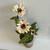 Three artificial zinnia flowers with cream petals speckled with pink and yellow centers, green leaves, and a small pot, set against a plain white wall.
